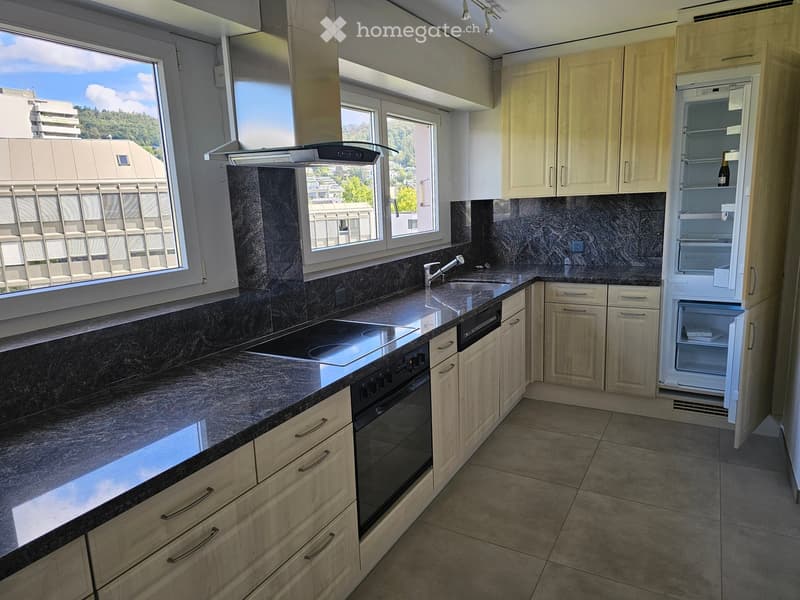 Modern kitchen with light wood cabinets, granite countertops, stainless steel sink, oven, and refrigerator. Large windows provide natural light and a view of the city.