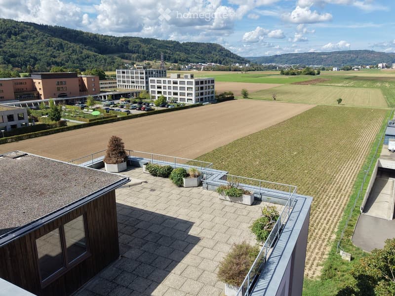 Aerial view of a modern building complex with a spacious rooftop terrace, surrounded by open fields and distant hills.