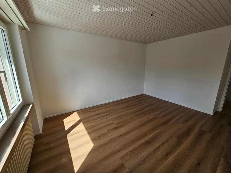Empty room with wooden flooring, white walls, and a window on the left side. The room has a slanted wooden ceiling and a radiator below the window.