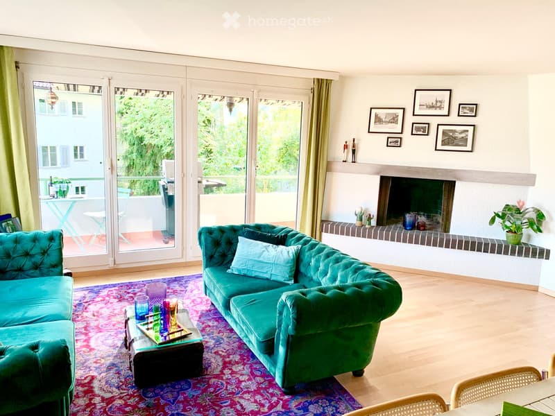 Modern living room with teal sofa, glass coffee table, fireplace, large windows, and outdoor seating area visible through glass doors.