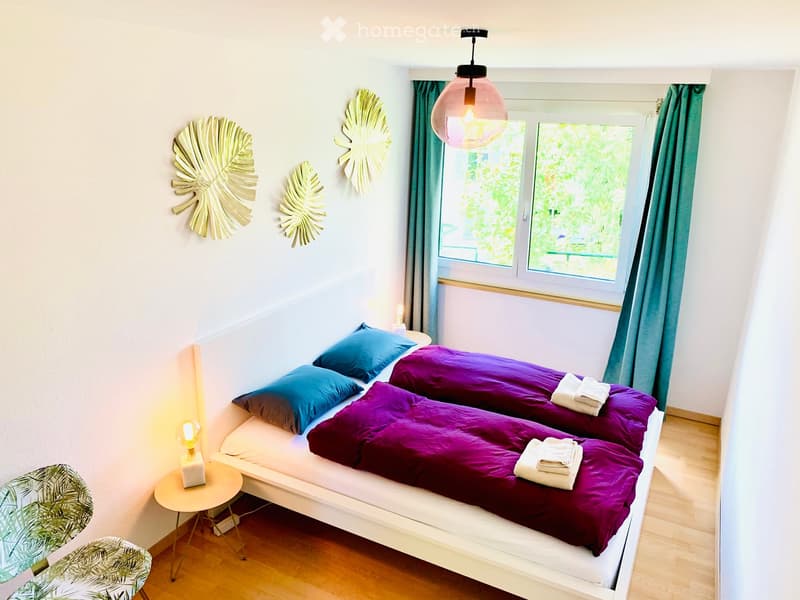 Cozy bedroom with two single beds, purple bedding, blue pillows, a small round table with a lamp, and decorative wall art.