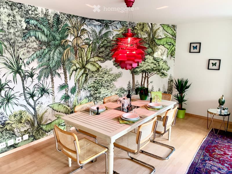 Dining area with a large table surrounded by chairs, set with plates, glasses, and a bottle. A colorful chandelier hangs above, and a large mural of a tropical scene adorns one wall. A small side table with plants and a patterned rug are also visible.