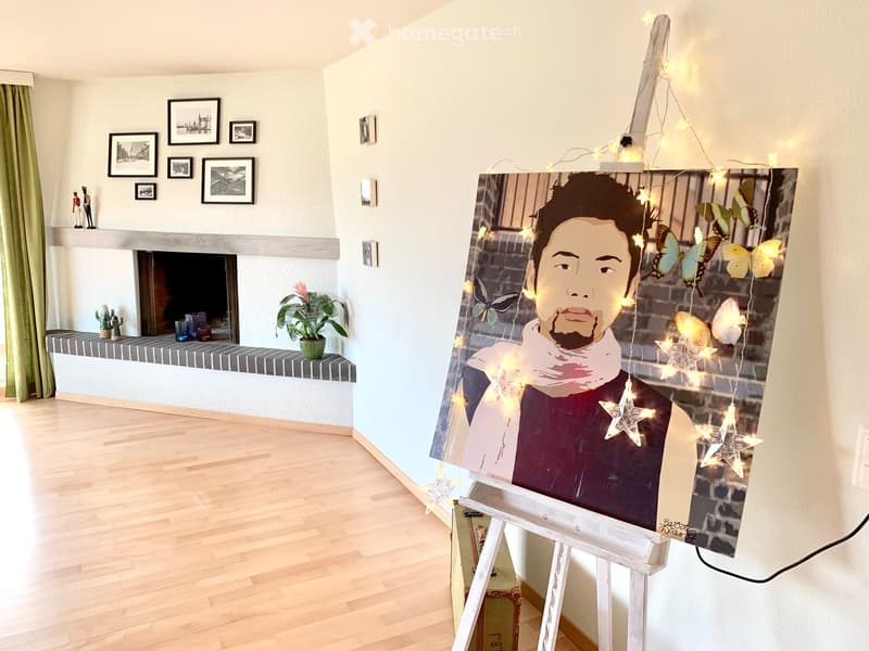 Living room with a fireplace, wooden floor, large painting on an easel, and decorative items on a shelf.