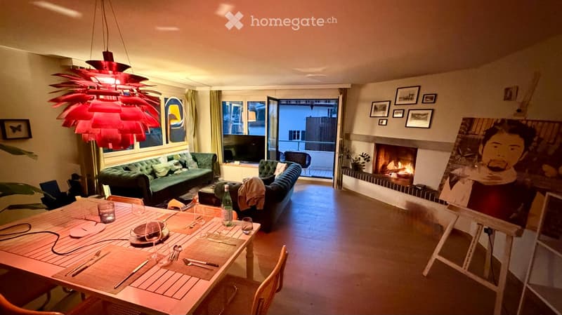 A modern living room with a green sofa, a dining table set for two, a fireplace, and large windows leading to a balcony.