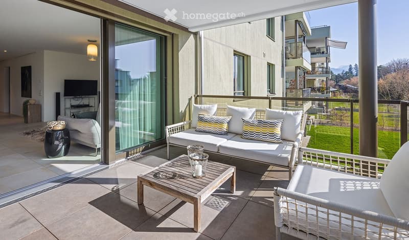 Balcony with a sofa, coffee table, and chairs, overlooking a green lawn and distant mountains.
