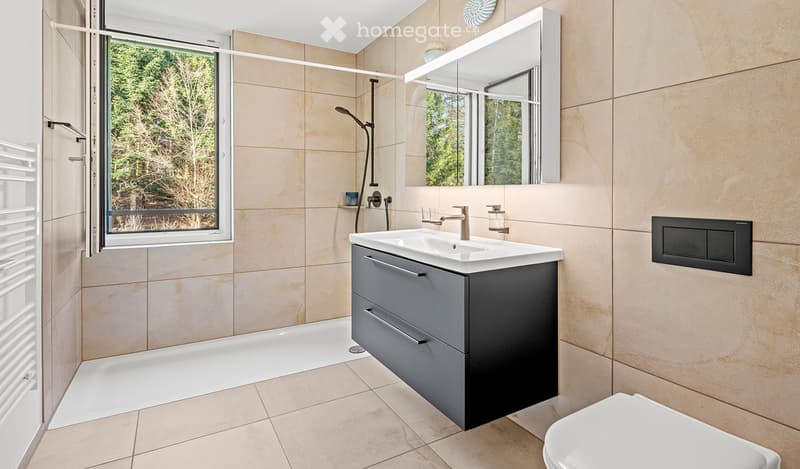 Modern bathroom with beige tiles, a large mirror, a sink with a modern faucet, a toilet, and a shower area with a glass door. The room features a radiator and a window with a view of greenery.