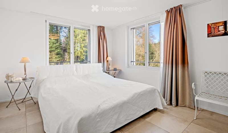 Bright bedroom with large windows, a neatly made bed, a nightstand with a lamp, a small table with a lamp, and a white bench near the window.