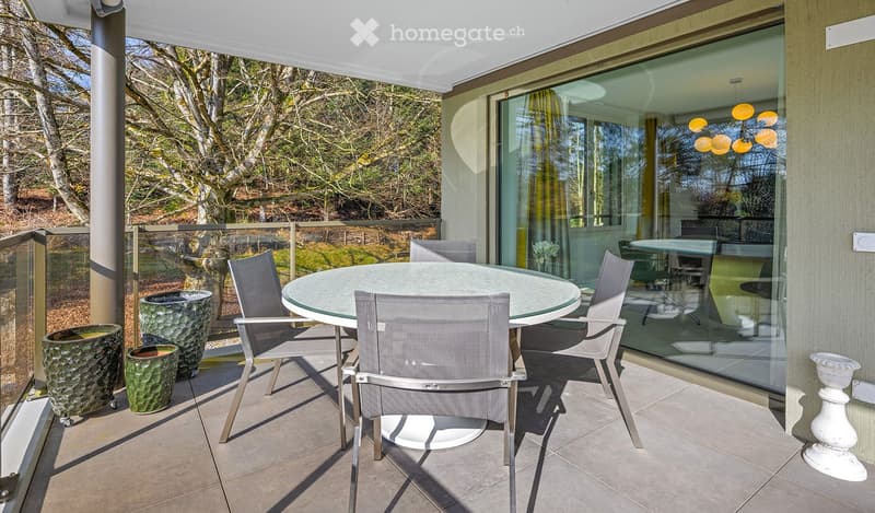 Balcony with a round glass table and four grey chairs, surrounded by large green planters and a view of a garden.