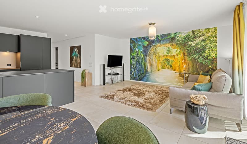 Modern living room with a large wall mural, a grey sofa with colorful cushions, a coffee table, a round dining table, and a TV stand with a flat-screen TV. The room features a sleek kitchen area with cabinets and a countertop.