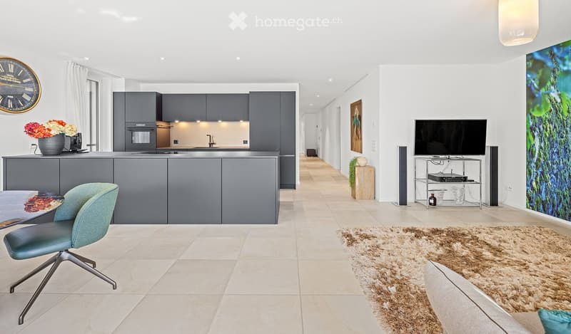 Modern open-plan living and dining area with a kitchen, featuring grey cabinets, a central island, a dining table with a teal chair, a television on a stand, and a large abstract painting on the wall.