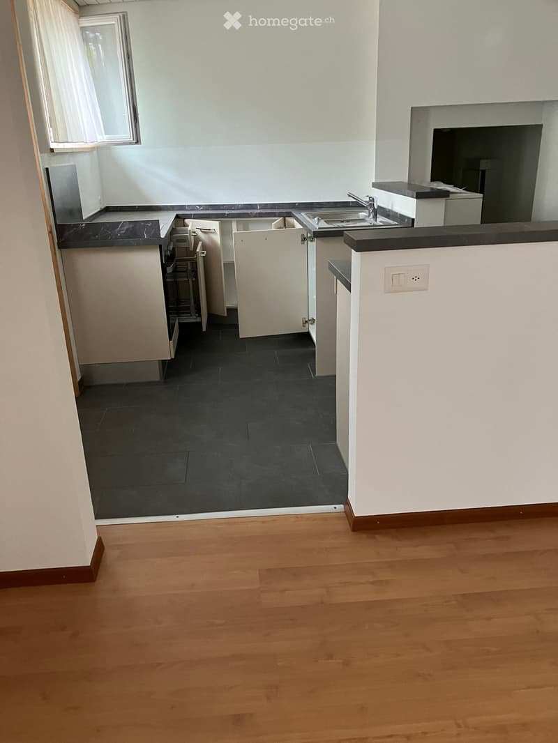 Empty room with wooden flooring and a small kitchen area featuring white cabinets, a sink, and a countertop. The kitchen area has dark countertops and a window with blinds.