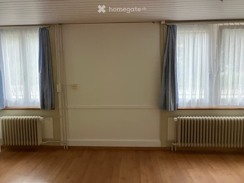 Empty room with wooden flooring, two windows with curtains, and radiators beneath each window.
