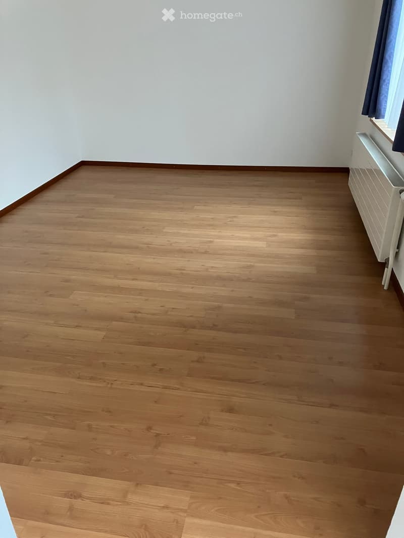 Empty room with wooden flooring and white walls, featuring a radiator and a window with blue curtains.