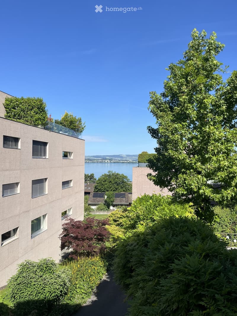 Modern building with a balcony and lush garden area, featuring various trees and shrubs, with a scenic lake view in the background.