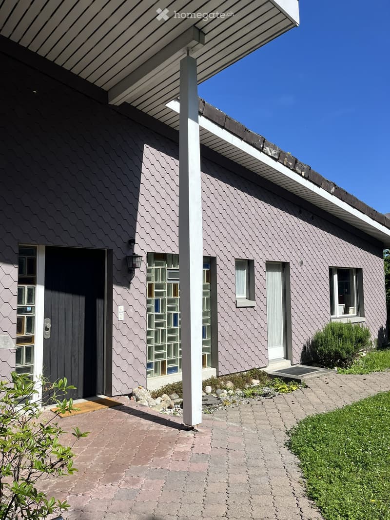 Single-story house with a modern, grey, textured facade, white trim, and a covered entrance supported by a white column. The entrance features a dark door with glass panels and a small doormat. The roof has a clean, white overhang. The surrounding area in