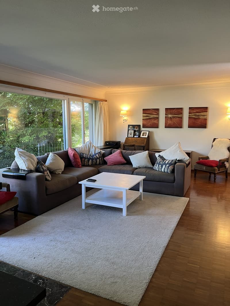 Modern living room with a grey sectional sofa, white coffee table, wooden floor, large windows with curtains, and artwork on the walls.