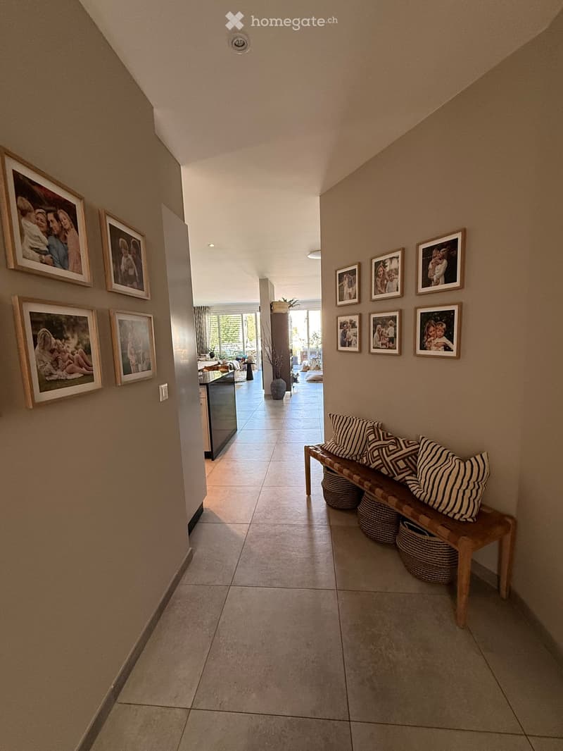 Hallway with beige tiled floor, wooden bench with striped cushions, framed family photos on walls, and a view into a living area with furniture and plants.