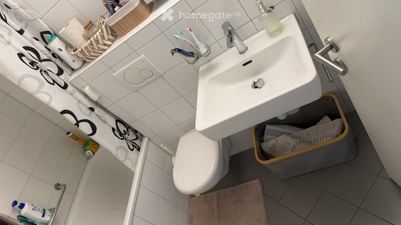 Modern bathroom with white tiles, a sink with a chrome faucet, a toilet, a shower curtain, a toothbrush holder with toothbrushes, a basket with toilet paper, and a trash bin.