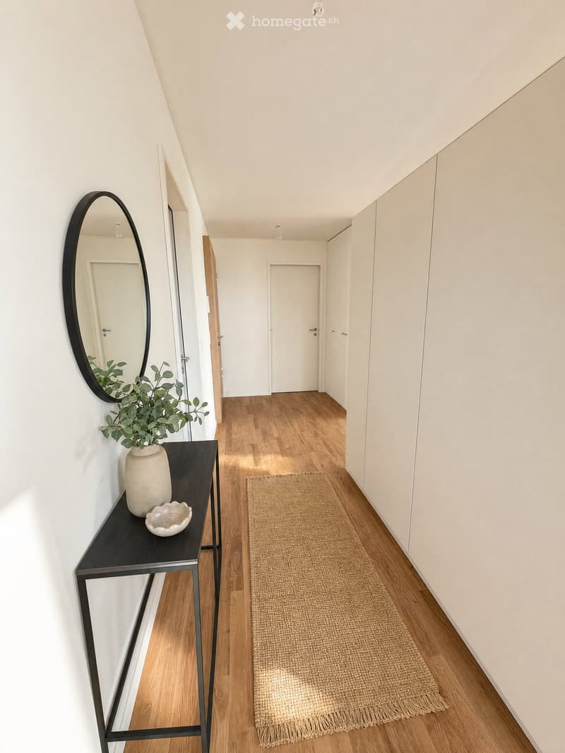 A modern hallway with wooden flooring, white walls, built-in wardrobes, a small black console table with a vase and bowl, a round mirror, and a natural fiber runner rug.