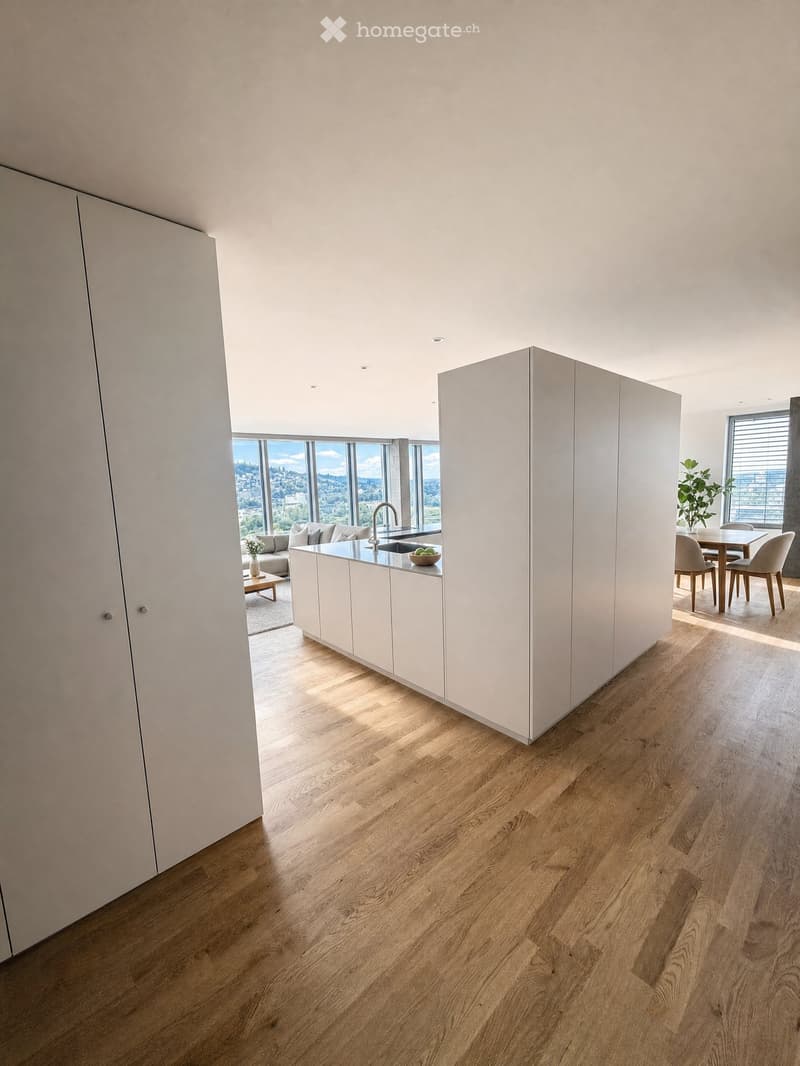 Open-plan living and dining area with modern white kitchen cabinets, wooden flooring, large windows offering a scenic view, and contemporary furniture including a sofa, coffee table, dining table, and chairs.