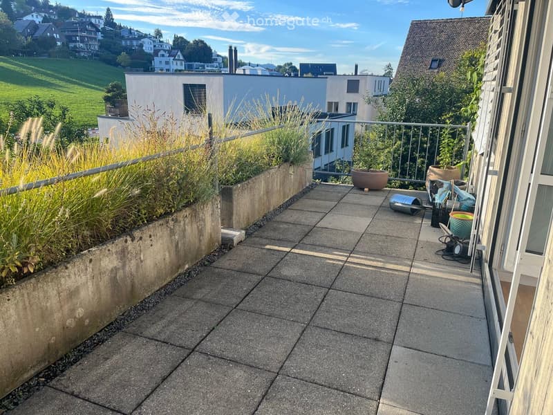Balcony with tiled floor, metal railing, potted plants, and a view of a green landscape and buildings.