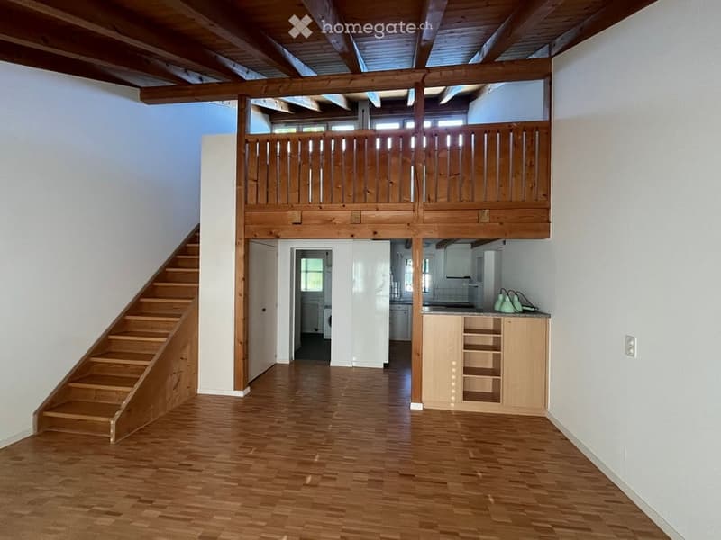 Open-plan living space with wooden flooring, exposed wooden beams, staircase leading to a wooden balcony, and a small kitchen area with cabinets.