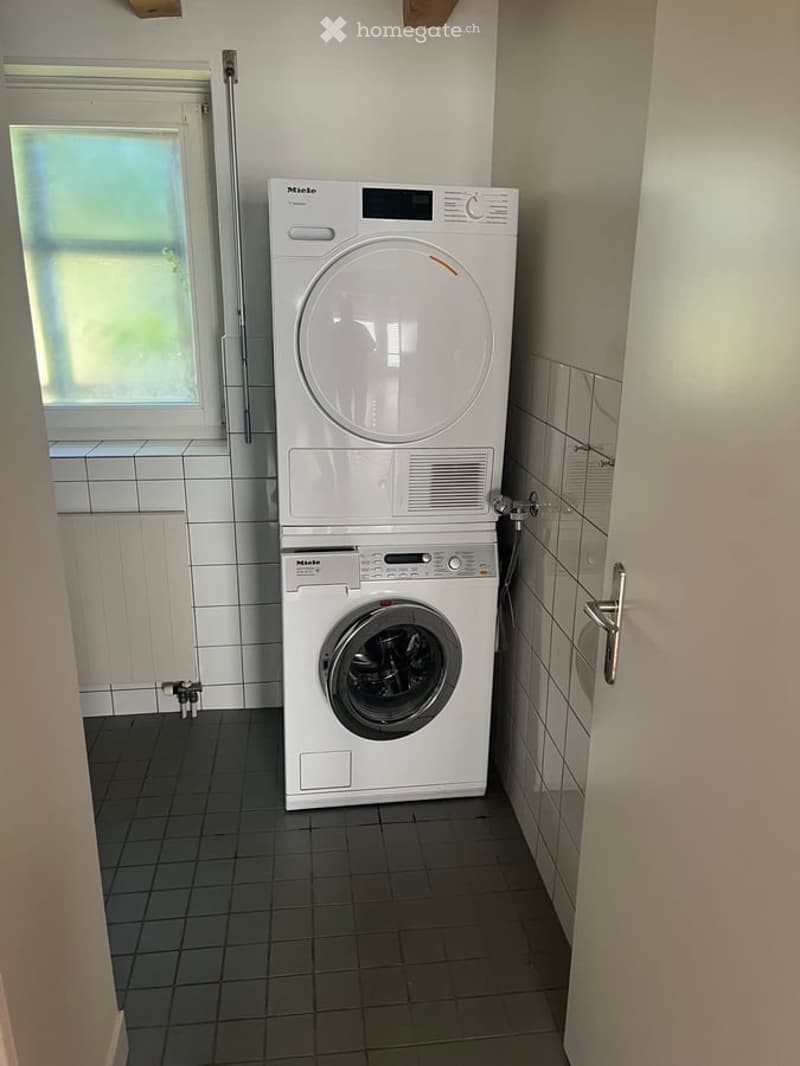 A compact bathroom with a stacked washing machine and dryer unit, white tiled walls, and a window providing natural light.