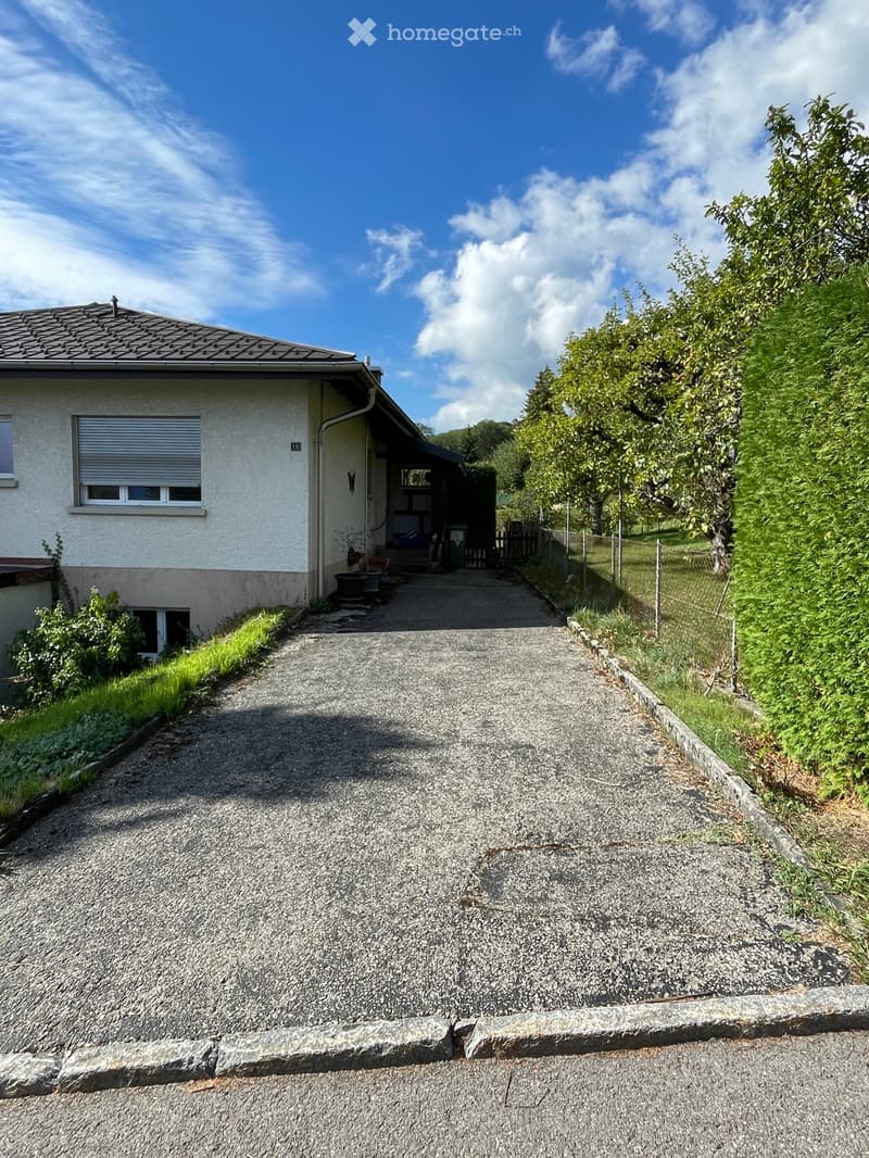 Single-story house with a gravel driveway, surrounded by greenery and trees. The house has a white facade, a grey roof, and a small porch area with a bench and potted plants.
