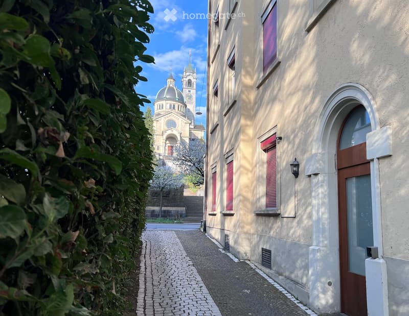 A narrow street with a brick pavement, a building with red shutters and a lamp on the wall, a church with a dome and a tower in the background, and a tree with white flowers.