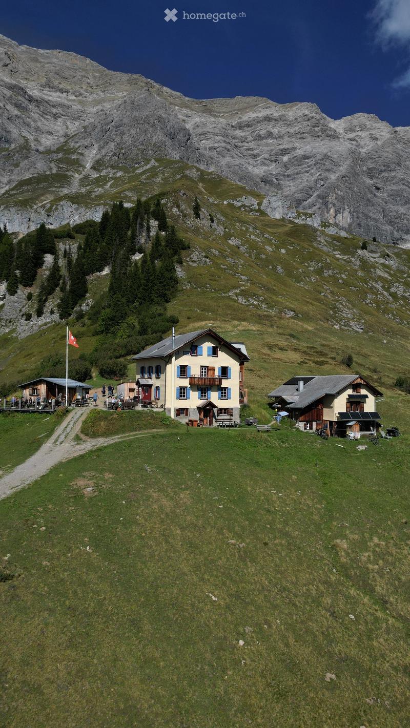 A cluster of small, yellow-painted houses with blue shutters and balconies, situated on a grassy slope surrounded by mountains and trees.