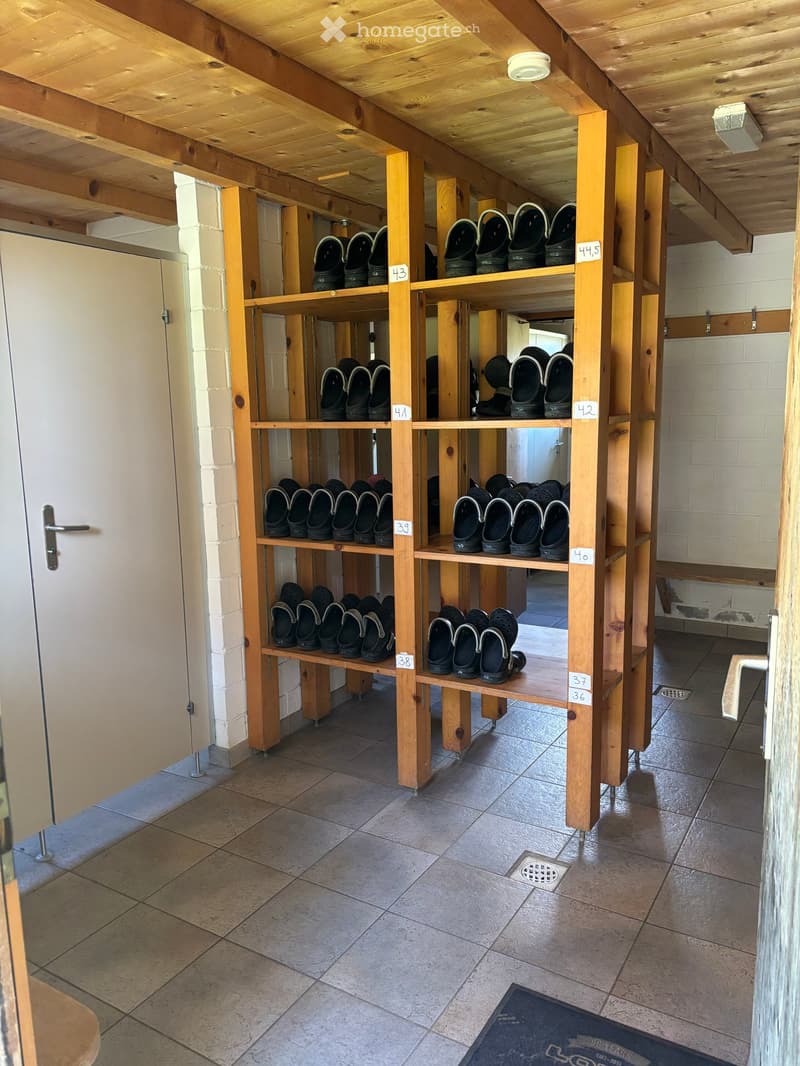Wooden shoe racks with numbered compartments holding black slippers, tiled floor, white brick walls, and a partially visible door.
