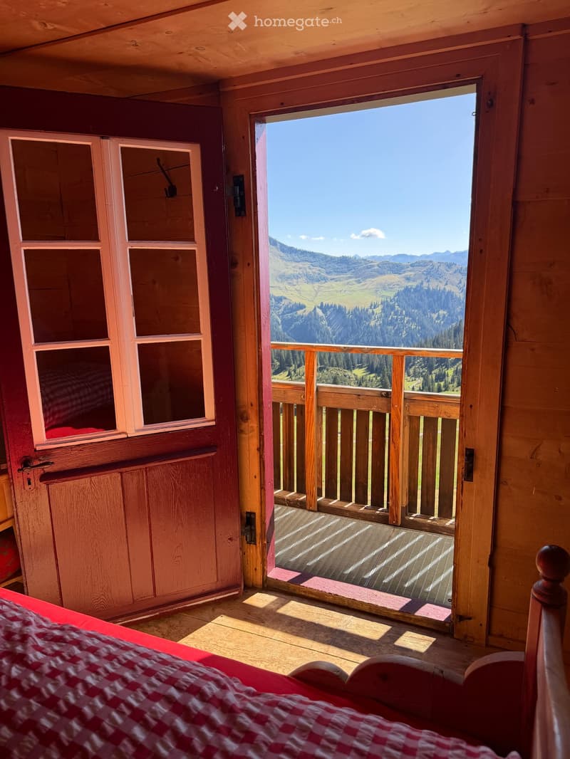 Cozy wooden bedroom with a large window offering a scenic mountain view, featuring a red and white checkered bedspread and wooden furniture.