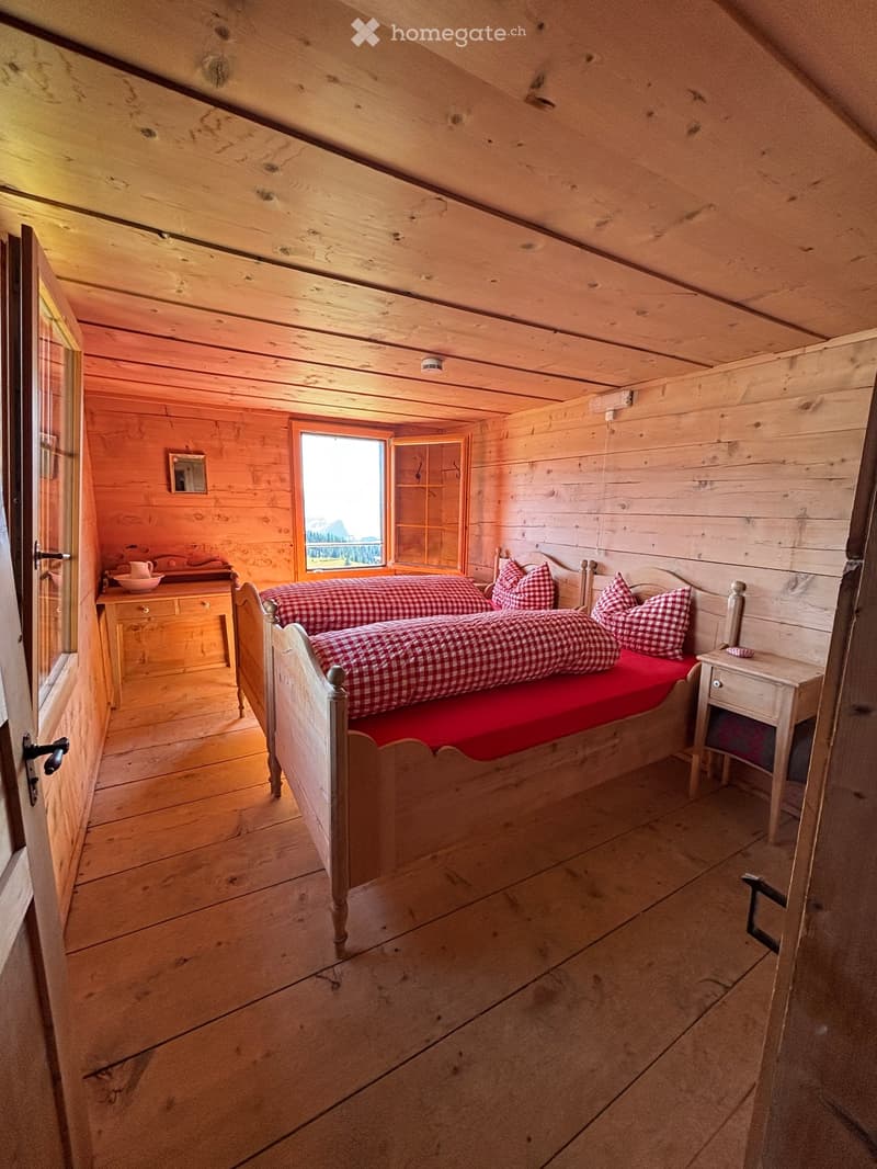 Cozy wooden bedroom with slanted ceiling, featuring a double bed with red and white checkered bedding, a wooden nightstand, a small mirror, and a wooden cupboard with a teacup on top.