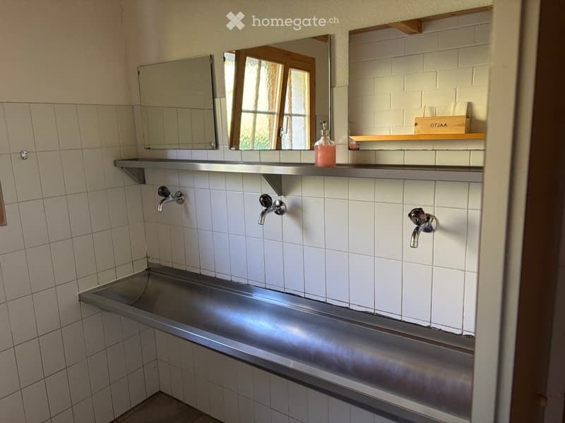 Bathroom with tiled walls, stainless steel sink, multiple faucets, mirrors, and a small shelf holding a soap dispenser and other items.