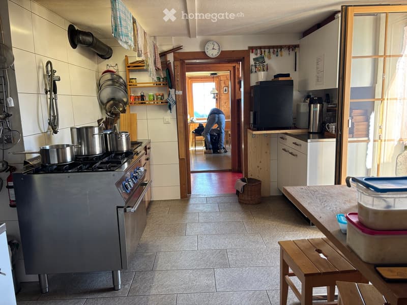The kitchen features a stove with pots, a refrigerator, a wooden table with a stool, a clock on the wall, and a doorway leading to another room.