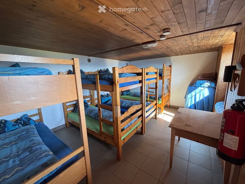 A cozy wooden bunk bed room with multiple bunk beds, a small wooden table, and a storage cabinet. The room features a wooden ceiling and tiled floor.