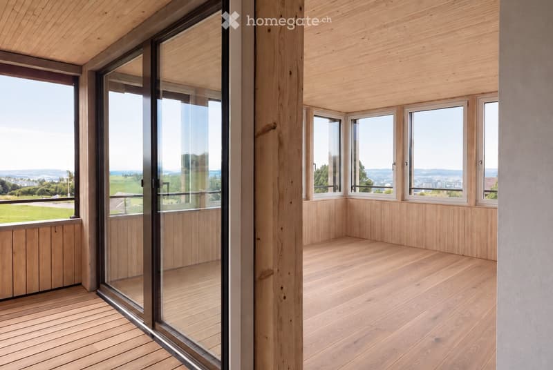 Modern wooden balcony with large glass sliding doors, expansive view of green fields and distant cityscape.