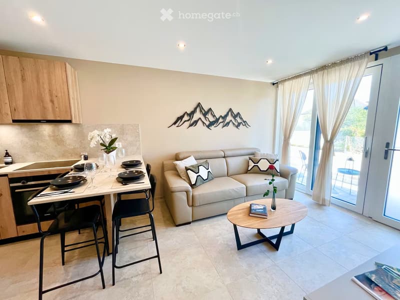Furnished Room. Modern open-plan living and dining area with a cream sofa, coffee table, dining table with stools, kitchen area with cabinets and appliances, and decorative wall art.