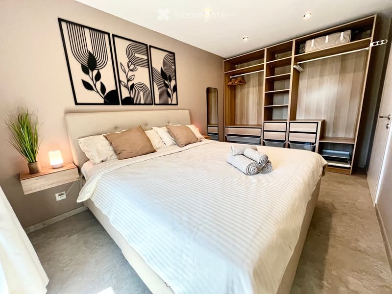 Bedroom. Modern bedroom with a large bed, nightstand, lamp, mirror, and built-in wardrobe with drawers. Features decorative wall art and a potted plant on the nightstand.