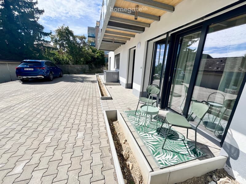 Balcony with modern furniture including chairs and a small table, surrounded by glass walls and a paved walkway.