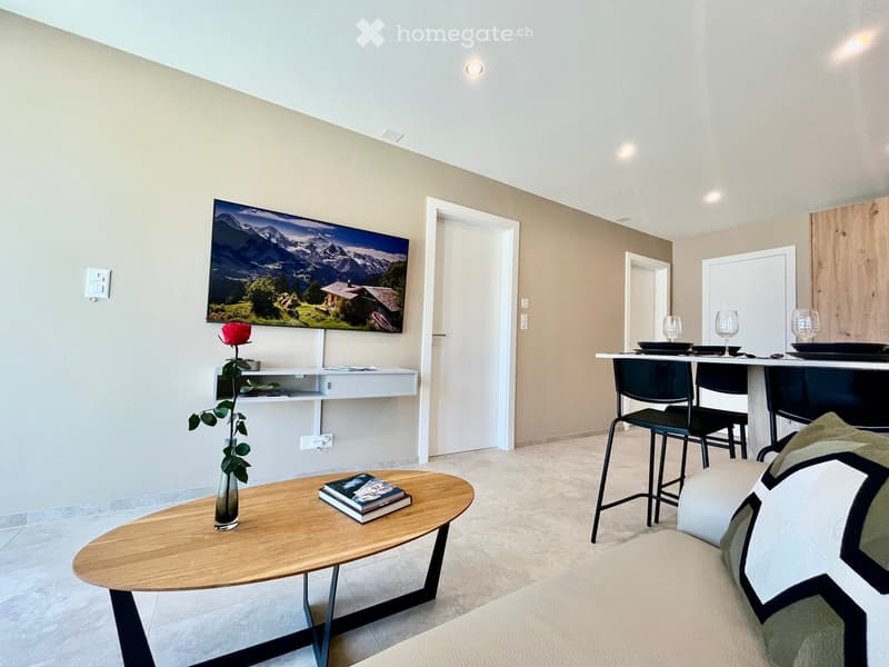 Modern living room with a sofa, coffee table, and a small bar area with stools. A large screen displays a scenic mountain view, and a vase with a red rose sits on a shelf.