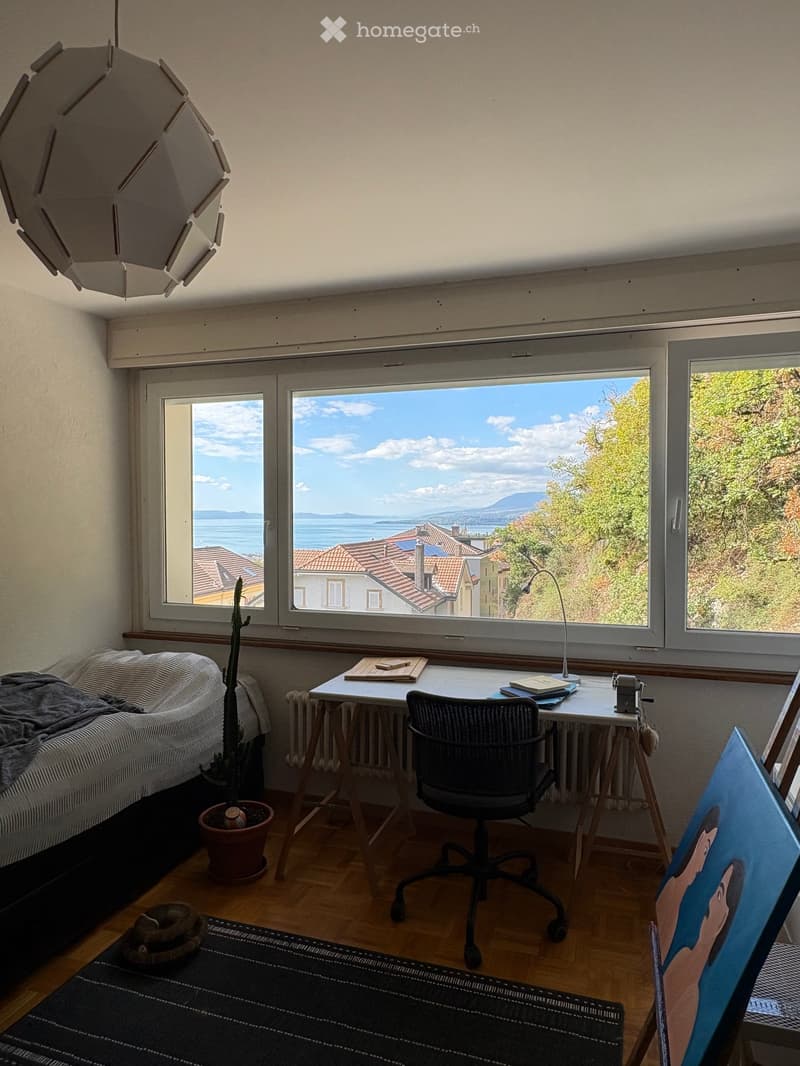 Bright room with large windows offering a scenic view, furnished with a sofa, desk, chair, lamp, and a painting on the wall.