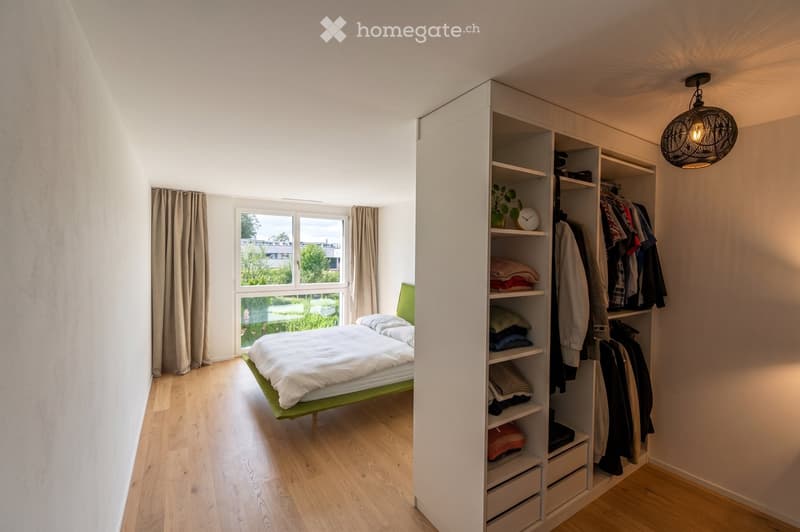 Modern bedroom with a green bed frame, white bedding, large window with curtains, built-in wardrobe with shelves and hanging space, and a hanging light fixture.