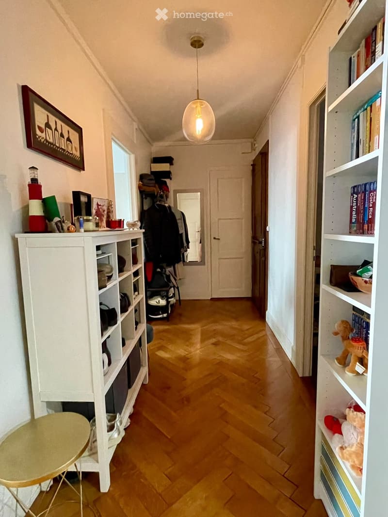 A narrow hallway with wooden flooring, white walls, and built-in shelving units on both sides. The left side features a small round table, a coat rack with jackets, and a framed picture. The right side has bookshelves with books, toys, and decorative item
