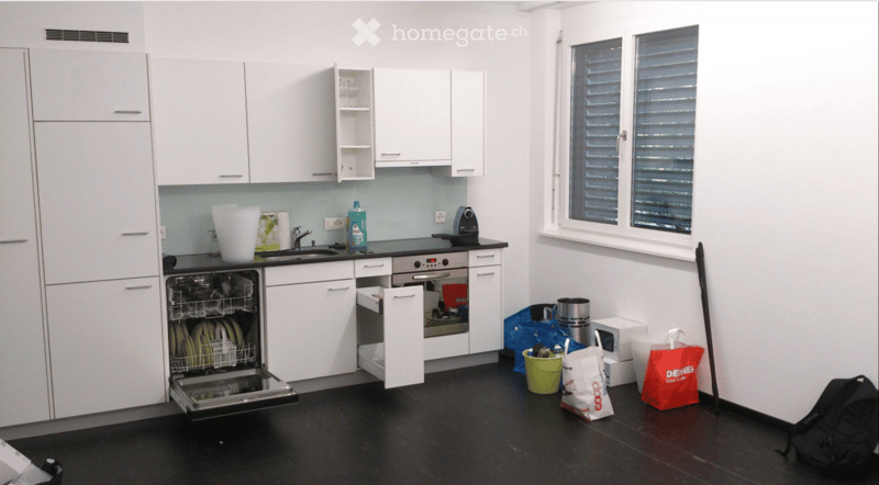 Modern kitchen with white cabinets, black countertops, and stainless steel appliances. Features a dishwasher, oven, sink, and various items including a backpack and handbags.