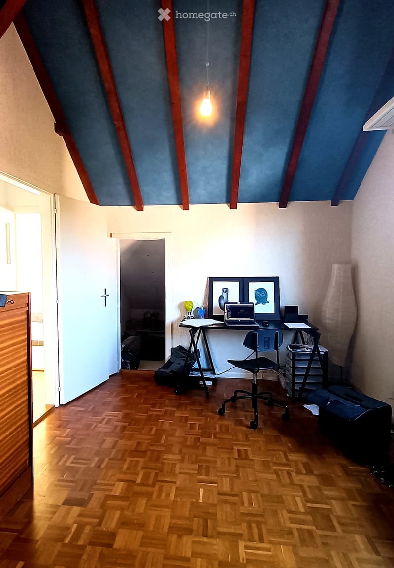 A modern, furnished room with wooden flooring, a desk with a laptop, a chair, and framed artwork on the wall. The room features exposed wooden beams and a slanted ceiling.