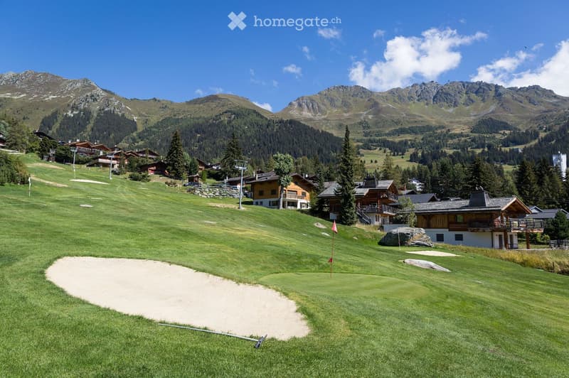 A scenic golf course surrounded by lush green grass, with several wooden chalets and houses in the background, set against a backdrop of snow-capped mountains and a clear blue sky.