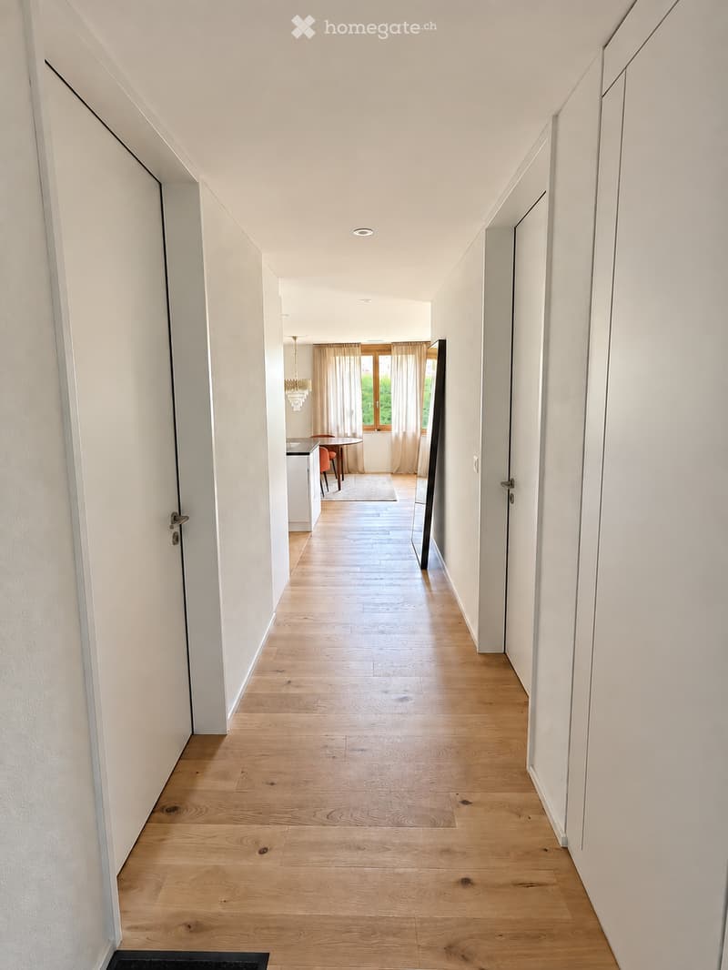 A long, narrow hallway with light wooden flooring and white walls, leading to a room with a dining table and chairs visible at the end.