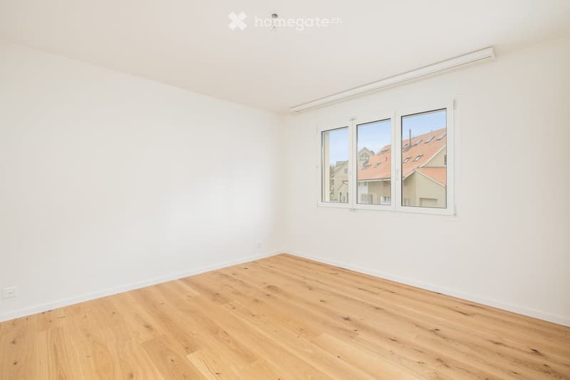 Empty room with white walls, wooden floor, and large windows showing a view of neighboring houses.