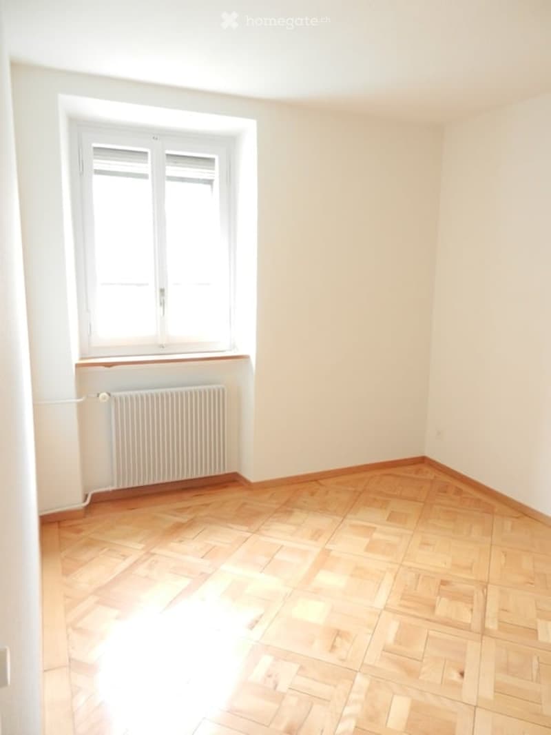 Empty room with wooden flooring, white walls, and a window with white frames. A radiator is visible below the window.